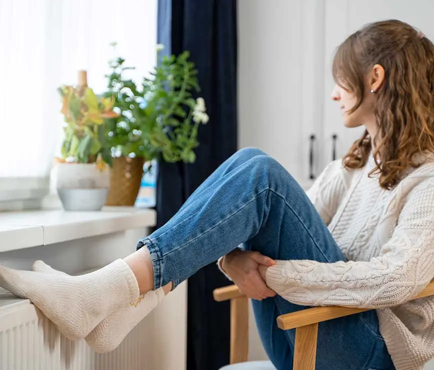 Frau sitzt gemütlich am Fenster mit Blick nach draußen und genießt die wohlige Wärme der Heizung.